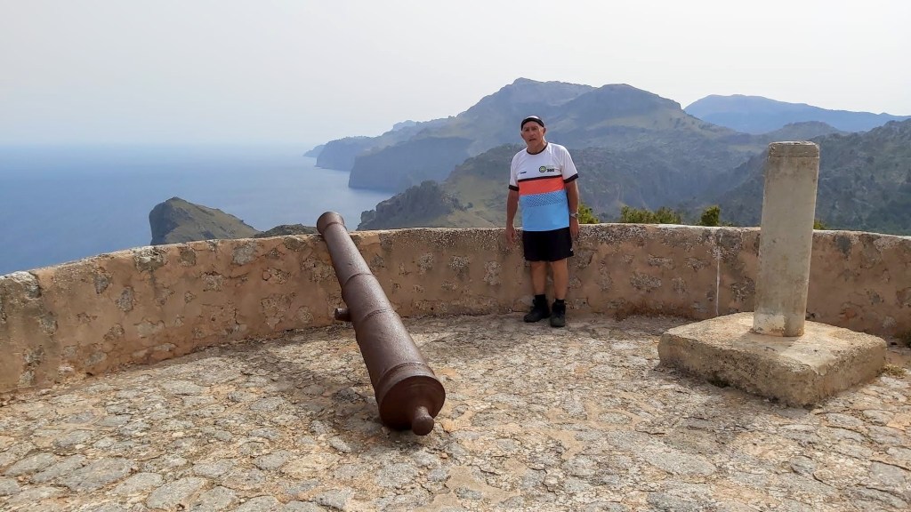 Un hombre de pie junto a un cañón en la Torre de la Mola de Tuent, con vistas panorámicas de montañas y costa al fondo.