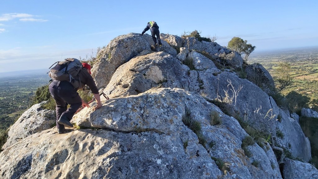 Serra de Galdent desde Llucmajor - Caminando por Mallorca Dos senderistas escalando una roca en el Puig de ses Bruixes, con vistas a un valle verde y un cielo despejado.
