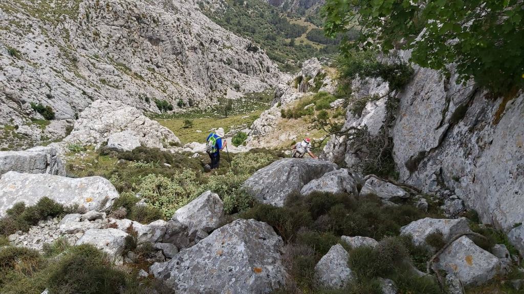 Vista de excursionistas en un terreno rocoso ascendiendo por un sendero entre grandes rocas y vegetación, rodeados de montañas.