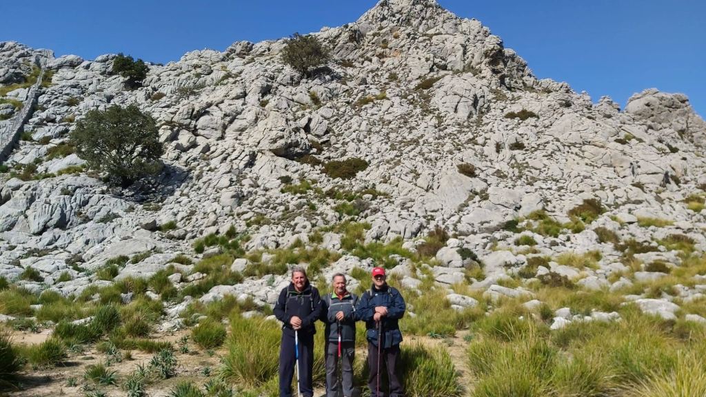 Tres senderistas en el camino hacia la Montaña de Moncaire, rodeados de rocas y vegetación, con un paisaje montañoso de fondo.