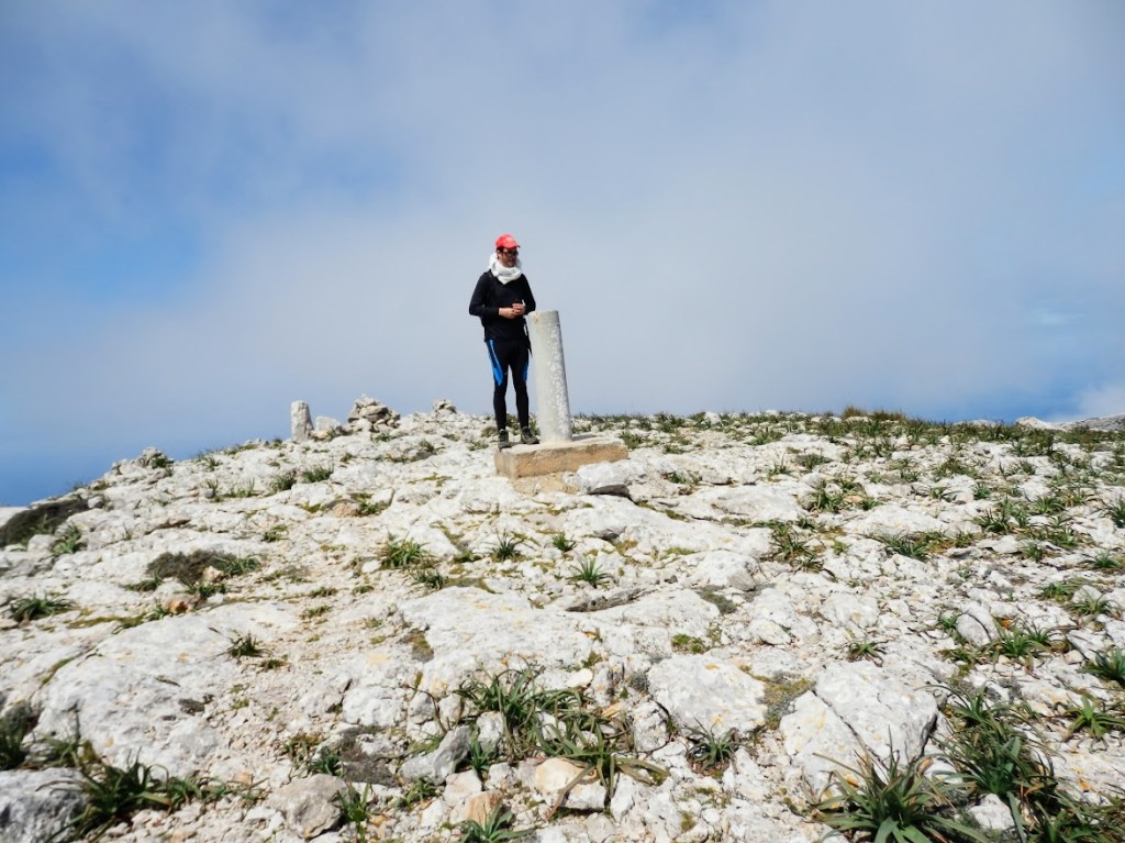 Persona de pie en la cima de la Mola de s'Esclop, junto a un vértice geodésico, rodeada de un paisaje rocoso y nuboso en la Sierra de Tramuntana.