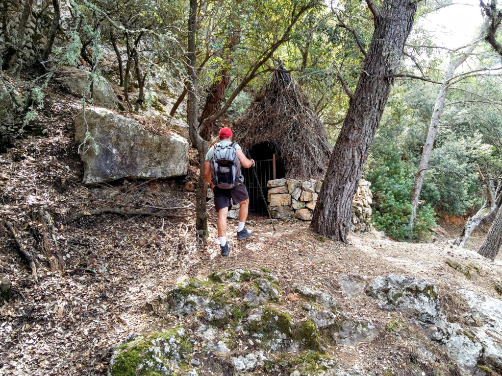 Un senderista se aproxima a una  barraca de carbonero habilitada para guardar utensilios de los cazadores, rodeada de vegetación en un bosque.
