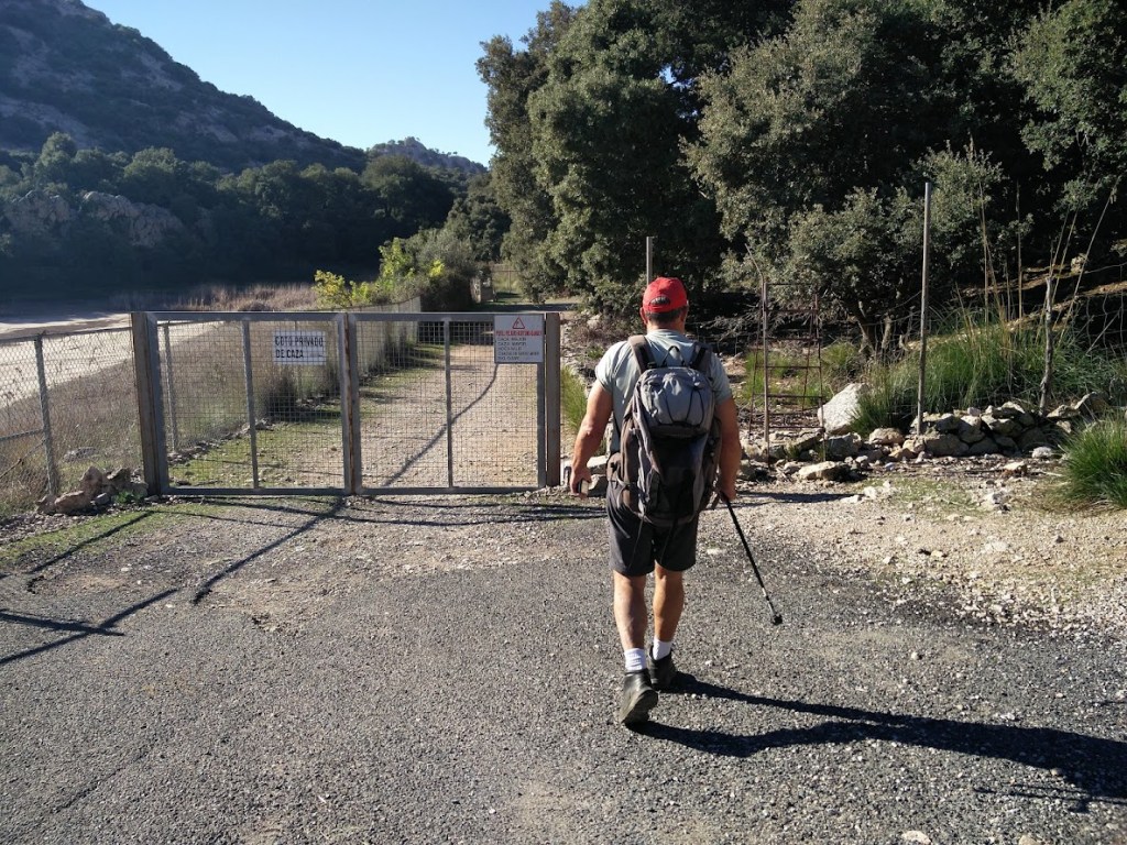 Senderista caminando hacia una puerta de acceso en un camino rural, rodeado de vegetación y montaña en la Coma de Son Torrella.