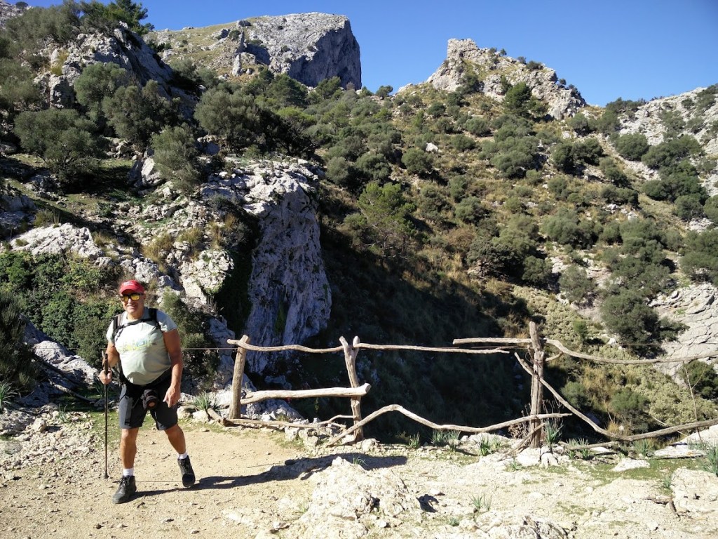 Senderista en un camino de montaña con un paisaje rocoso y vegetación en el fondo, sosteniendo un bastón de trekking.