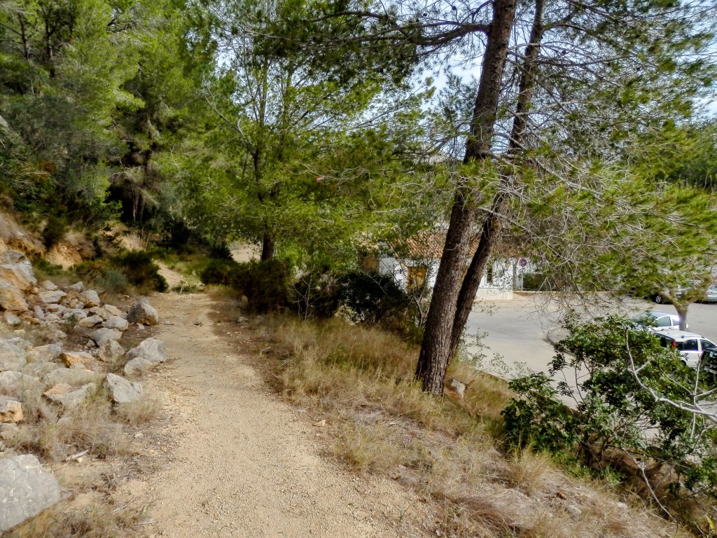 Sendero de tierra rodeado de vegetación y árboles, con el aparcamiento del Cementerio de s’Arracó.