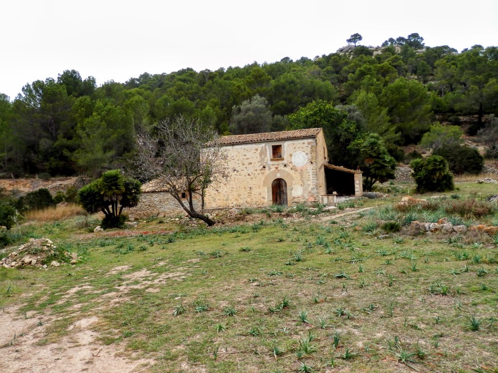 Vista de una antigua construcción rural en un entorno natural, rodeada de vegetación y árboles. La edificación presenta una fachada de piedra con detalles arquitectónicos simples y un paisaje montañoso al fondo.