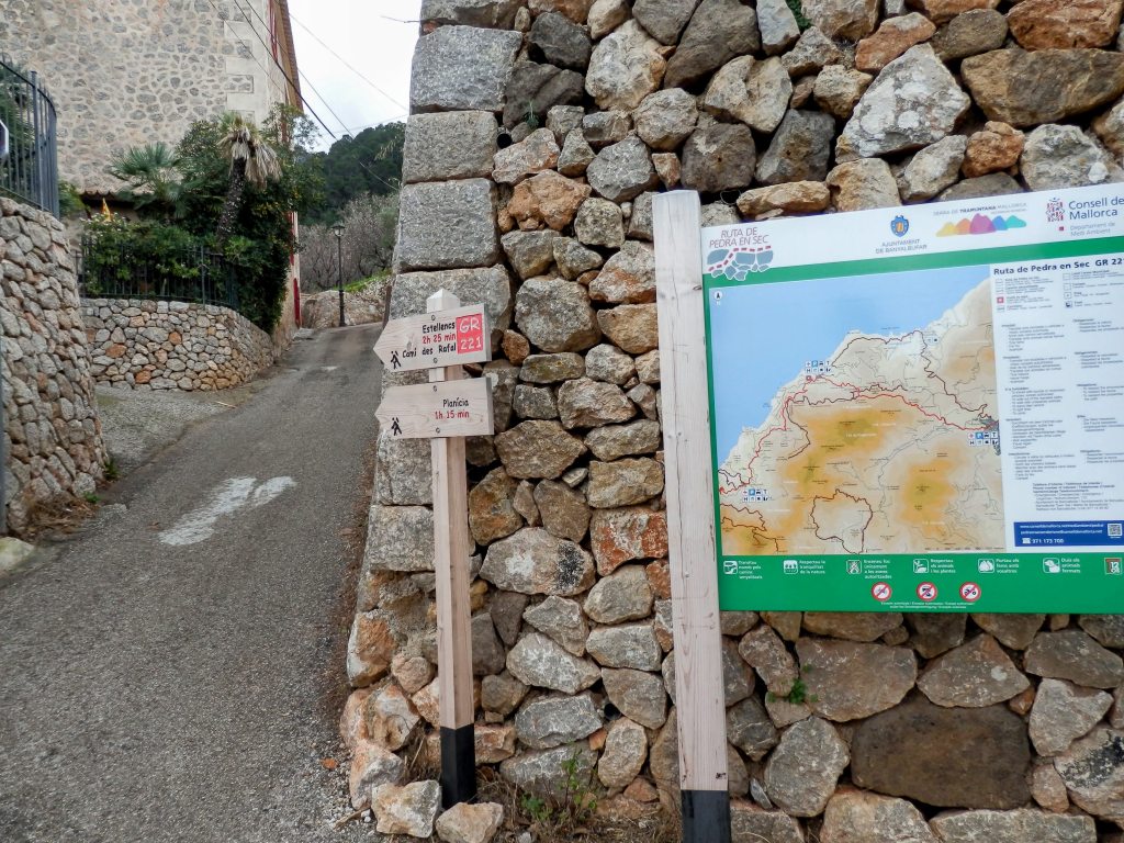 Señalización de la ruta GR 221 hacia Estellencs y Planícia, junto a una pared de piedra en un camino de montaña.