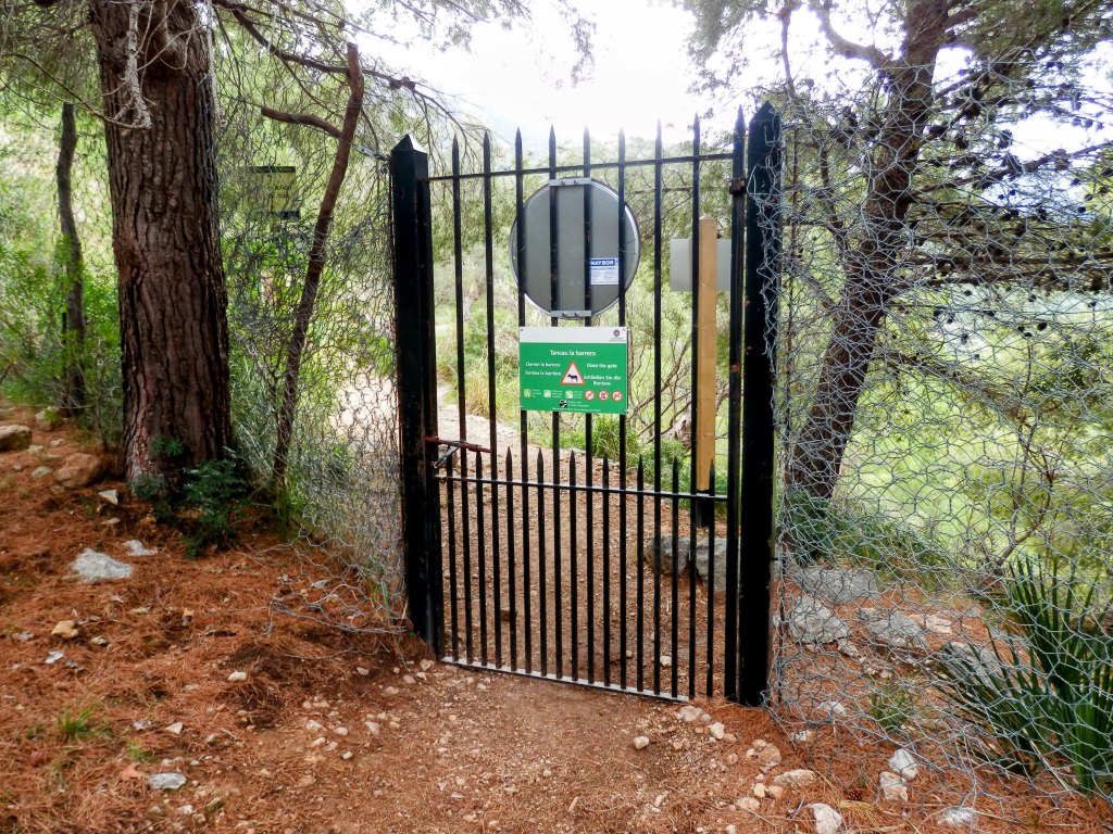 Puerta de acceso con reja negra y cartel informativo en un entorno natural con árboles.