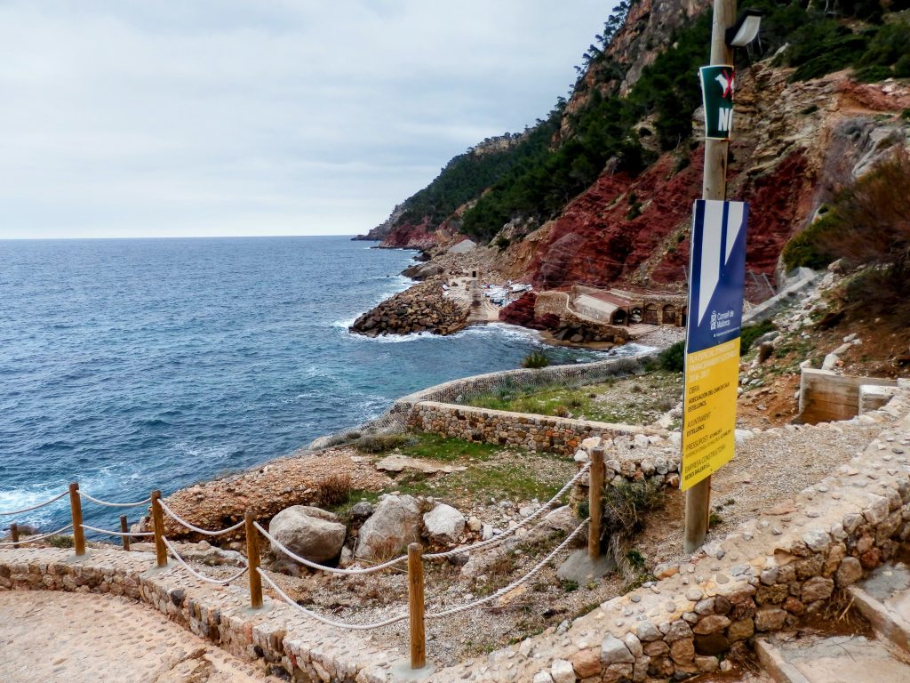 Vista de la Cala d'Estellencs con aguas azules y acantilados rocosos, junto a un sendero y un cartel indicador en la costa.