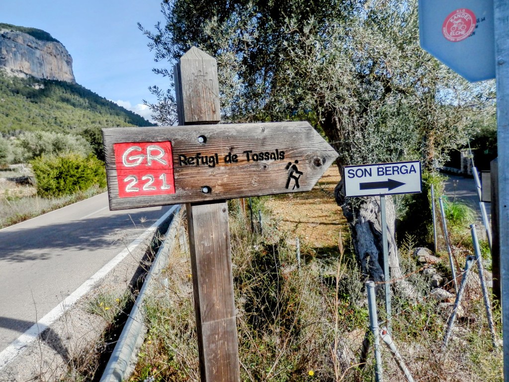 Señalización del GR 221 indicando la dirección hacia el refugio de Tossals Verds y Son Berga, con un fondo de paisaje montañoso.
