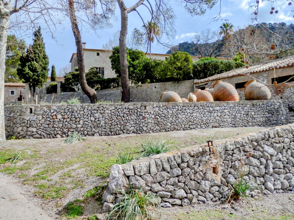 Vista de un entorno rural con un muro de piedra, árboles y jarrones de cerámica en el suelo, en el fondo se ven las casas de Solleric.