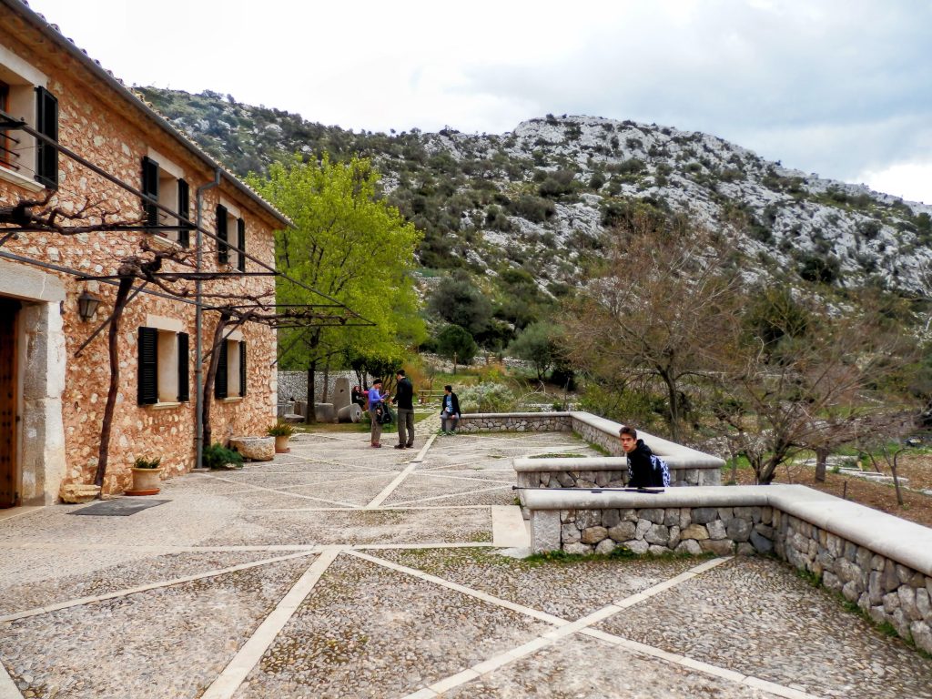 Vista del refugio de Tossals Verds, con su arquitectura de piedra, rodeado de un paisaje montañoso y vegetación. Se observa un patio empedrado donde hay personas interaccionando y árboles en el entorno.