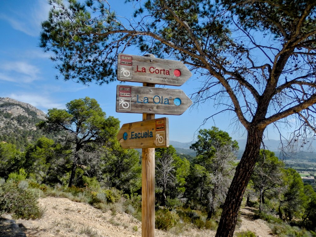 Señales de dirección de senderismo en una zona boscosa, indicando rutas como 'La Corta', 'La Ola', y 'Escuela', con un paisaje montañoso al fondo.