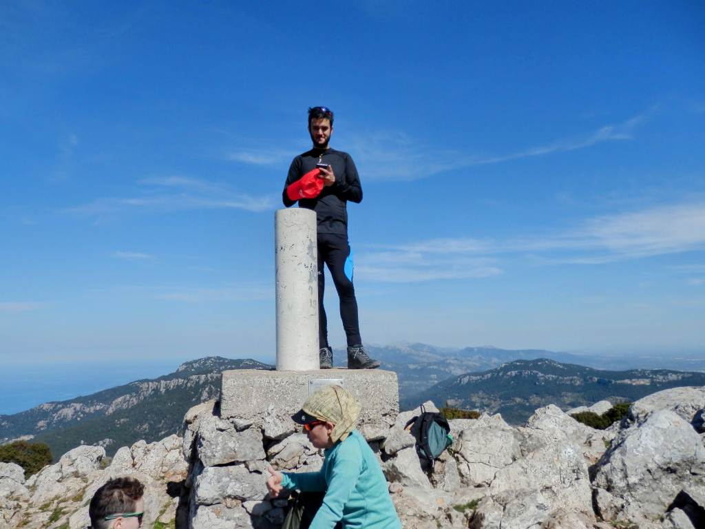 Persona de pie sobre un vértice geodésico en la cima del Puig des Galatzó, con el paisaje montañoso de fondo y un cielo despejado.