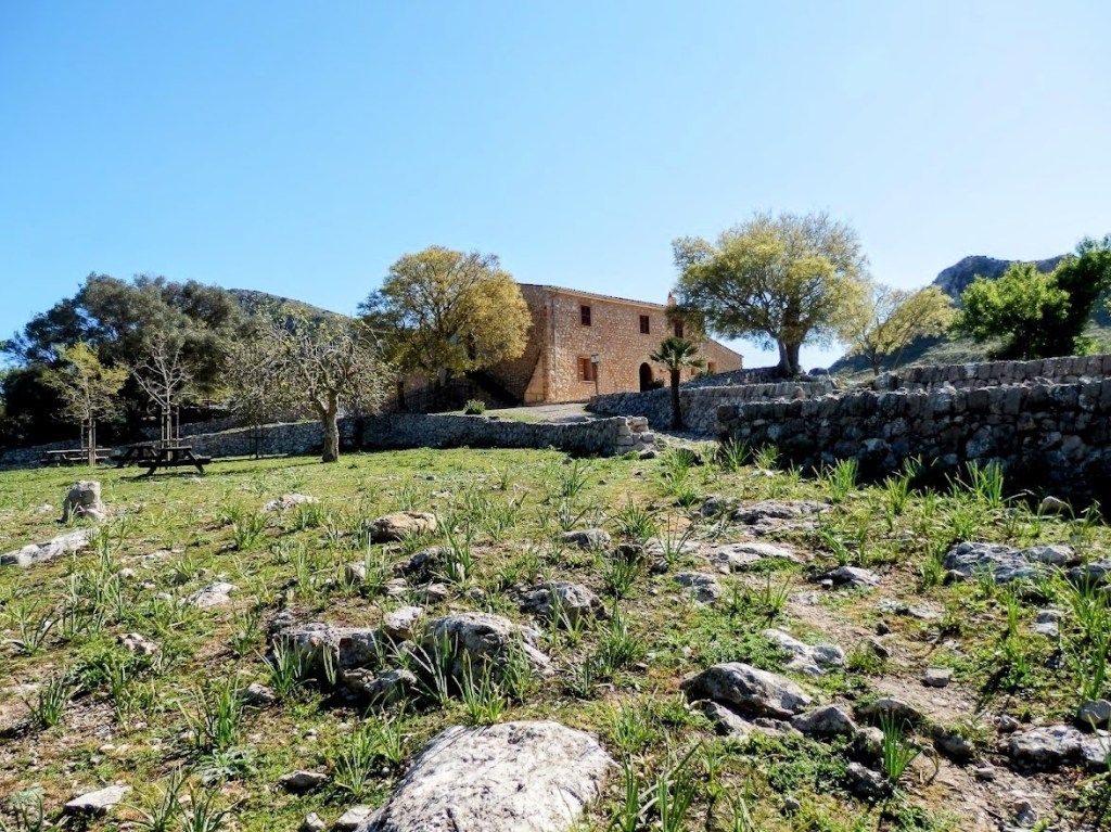 Vista del aparcamiento de s'Alqueria Vella de Baix, con un edificio de piedra y vegetación circundante en un día soleado.