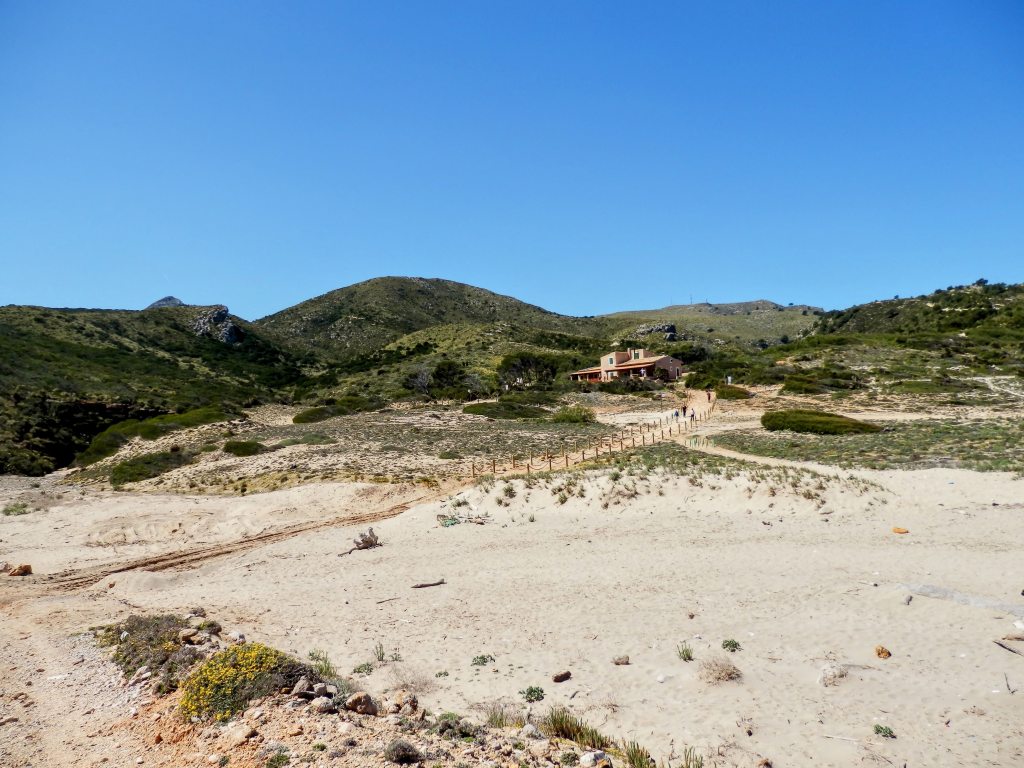 Vista de la playa S’Arenalet d’es Verger con el refugio de s’Arenalet al fondo, rodeado de vegetación y montañas bajo un cielo azul.