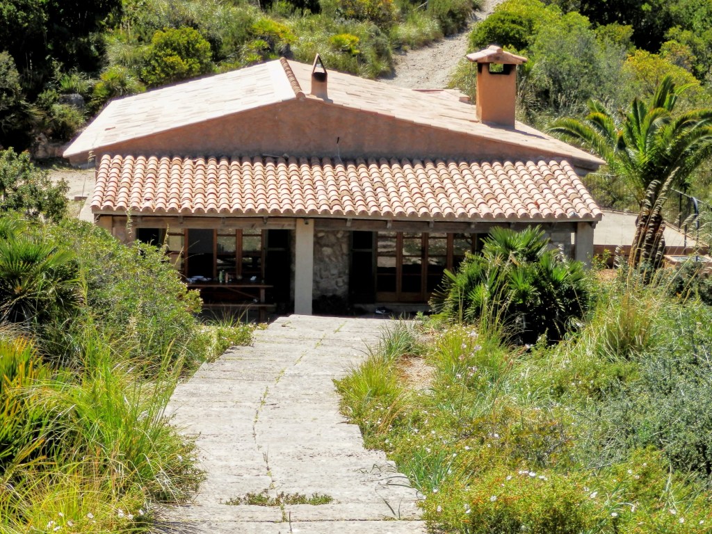 Vista del Refugio de s'Oguers, una construcción de una sola planta con una fachada de piedra y techo de tejas, rodeada de vegetación en un paisaje natural.