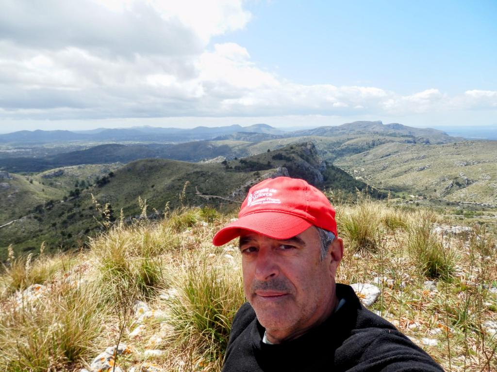 Un hombre con una gorra roja se toma un selfie en la cima del Puig des Porrassar, con un paisaje montañoso al fondo que muestra colinas y un cielo parcialmente nublado.