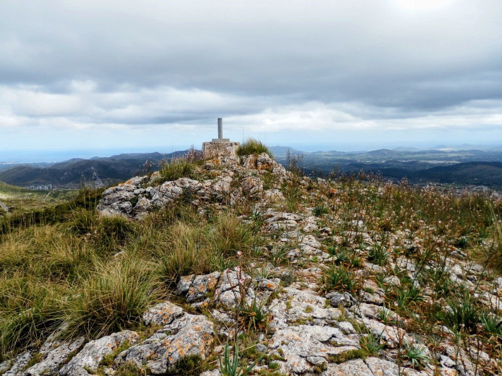 Vista panorámica desde la cima de Sa Talaia Freda, con vegetación y rocas, y un hito en el centro, con un cielo nublado de fondo.