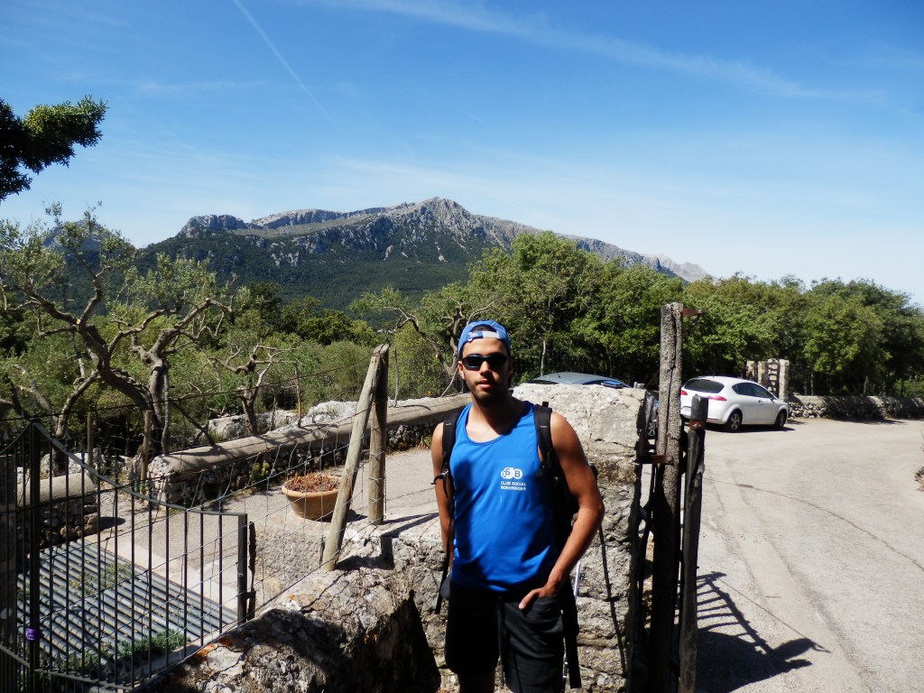 Hombre de pie frente a un paisaje montañoso, con árboles y una carretera de fondo, luciendo una camiseta sin mangas y una gorra.