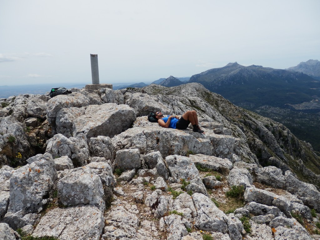 Persona descansando sobre rocas en la Cima del Puig Tomir, con un vértice geodésico visible en el fondo y vistas de las montañas y el paisaje circundante.