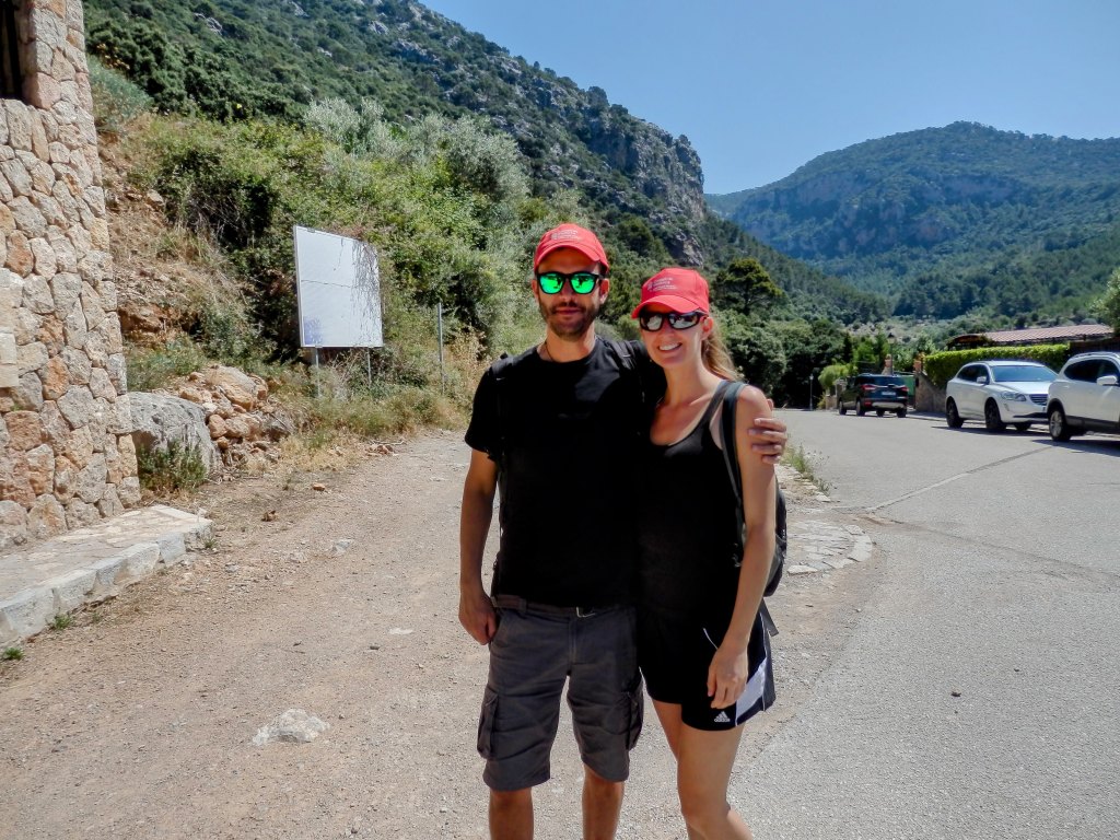 Una pareja sonriente de pie en un camino rural, rodeados de vegetación y montañas al fondo. Ambos llevan gorras rojas y gafas de sol. Se ven coches estacionados a la derecha en un día soleado.