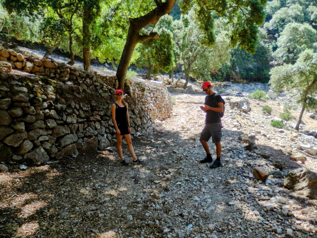 Dos personas caminando por un sendero rodeado de árboles y un muro de piedras en el paisaje natural.