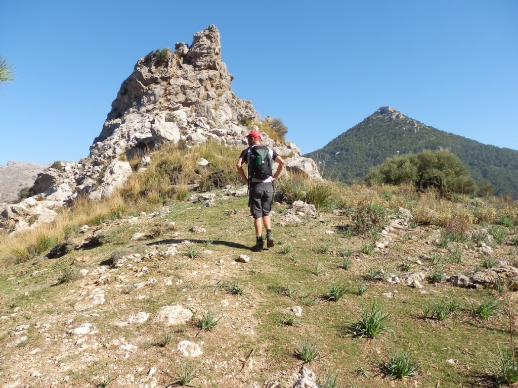 Persona caminando por un sendero rocoso en un paisaje montañoso, con formaciones rocosas al fondo y un cielo despejado.