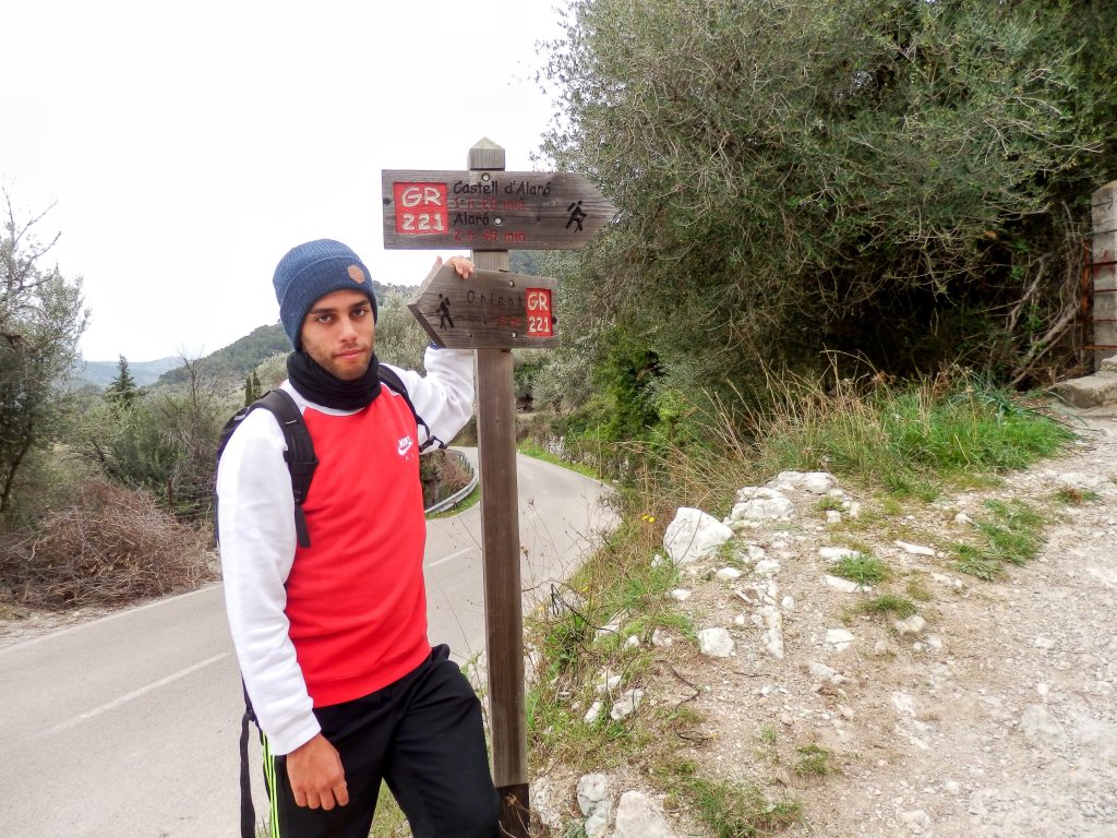 Joven en un sendero junto a un cartel indicativo del GR-221, señalizando la dirección hacia el Castell d'Alaró y Orient, con un paisaje montañoso al fondo.