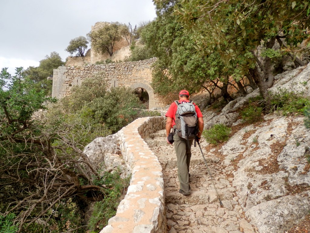 Sendero empedrado que lleva hacia las ruinas del Castillo de Alaró, con un excursionista caminando y vegetación circundante.