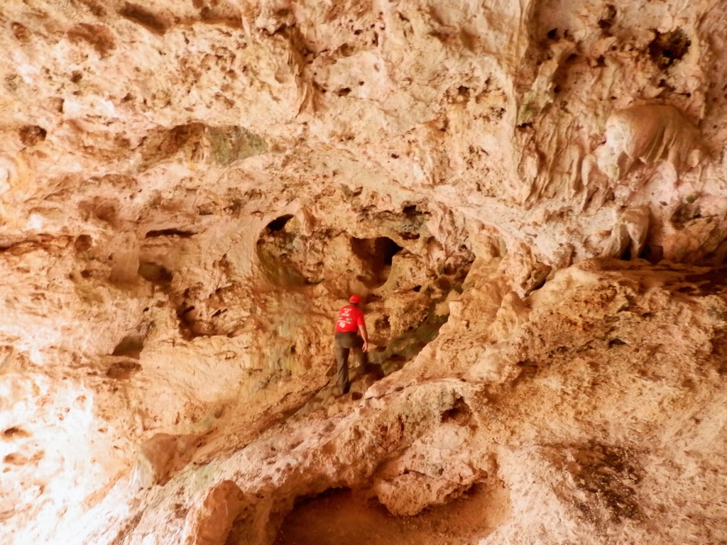 Interior de la Cova de Sant Antoni con un senderista explorando la cavidad y las formaciones rocosas en tonos beige y marrón.