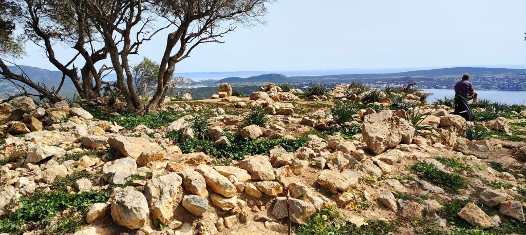 Vista panorámica desde la Cima del Castellot de na Marió, con rocas y vegetación en primer plano y un fondo que muestra el mar y las colinas.