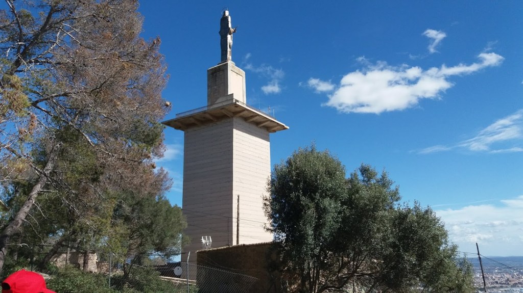 Vista del Monumento de Na Burguesa en el Puig de Na Burguesa, con una escultura en la cima y rodeado de árboles y cielo despejado.