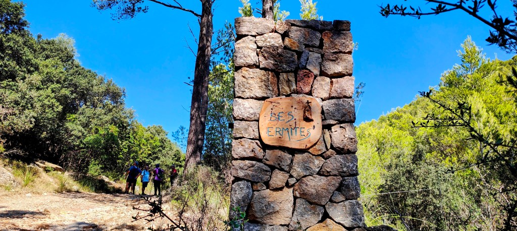 Señal de piedra que indica el camino hacia Ses Ermites, con un grupo de personas caminando por un sendero rodeado de vegetación.