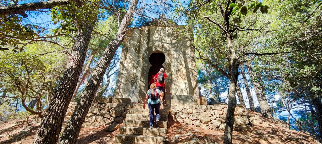 Dos personas subiendo las escaleras de  la Capella des Puig des Verger de estilo árabe, rodeadas de árboles en un entorno natural.