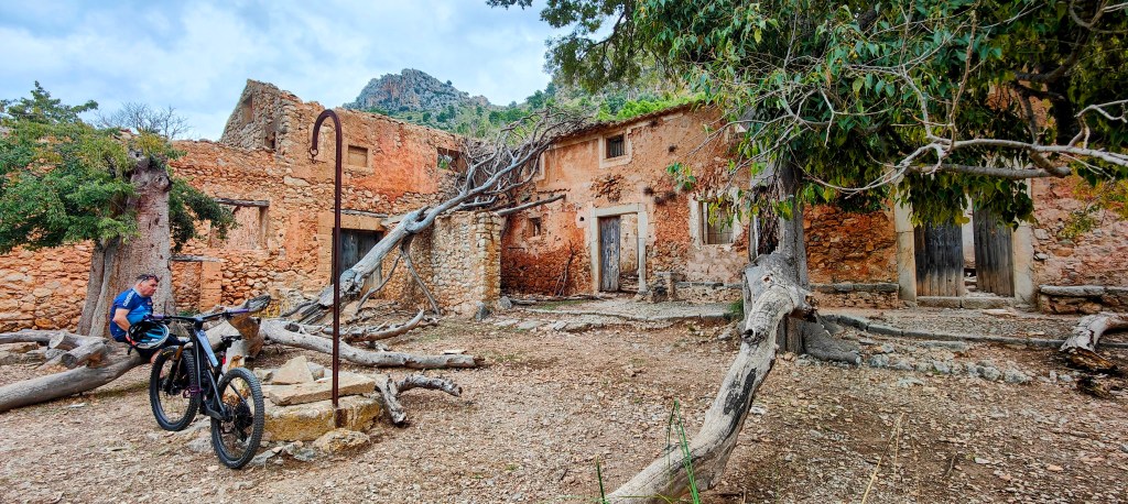 Un ciclista descansando junto a las ruinas de Raixeta, con árboles y montañas al fondo.