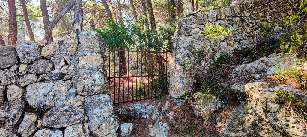 Entrada a un sendero natural con una puerta metálica roja y muros de piedra.