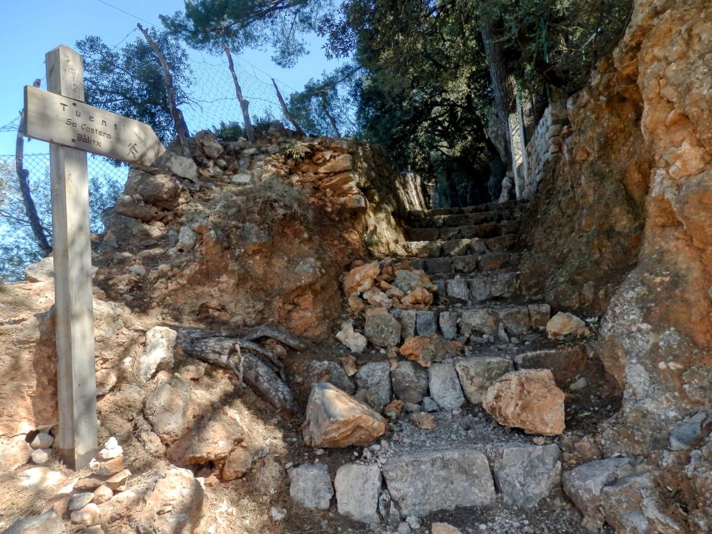 Escaleras de piedra en un sendero, con un cartel que indica direcciones hacia Tuent y Sa Costera, rodeado de vegetación y rocas.
