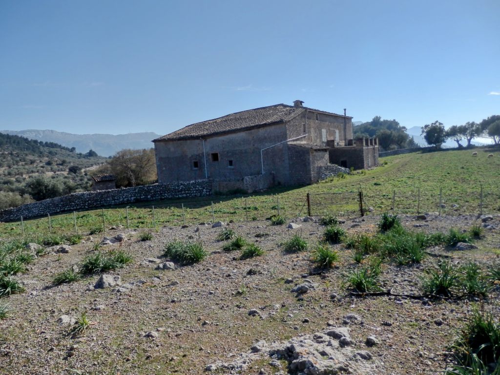 Vista de una antigua finca en Balitx d'Amunt, rodeada de terreno pedregoso y vegetación mediterránea, con montañas al fondo y un cielo despejado.