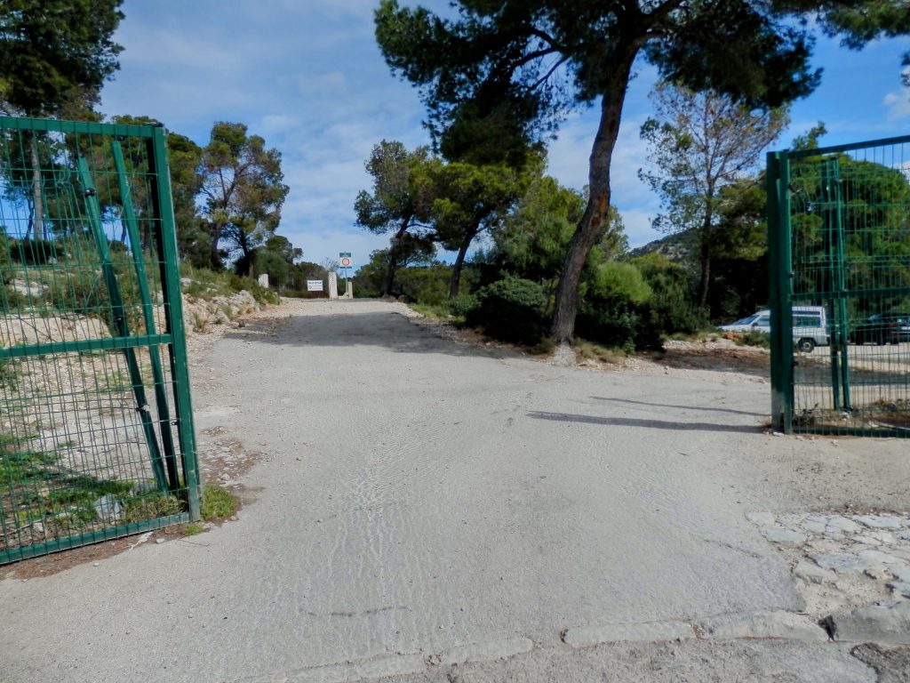 Entrada al camino de la ruta "Pi de Ses Creus" con una barrera verde a ambos lados, rodeado de árboles y un aparcamiento visible al fondo.