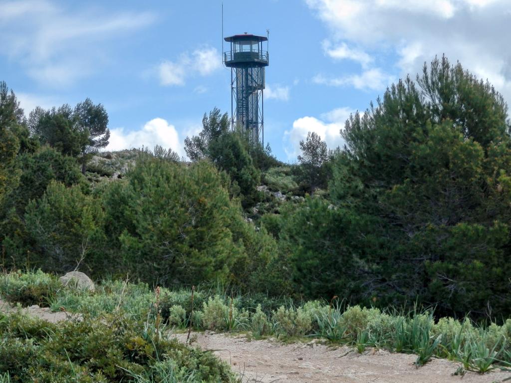 Torre de vigilancia contra incendios ubicada en el Pi de ses Creus, rodeada de árboles y vegetación, bajo un cielo con nubes.