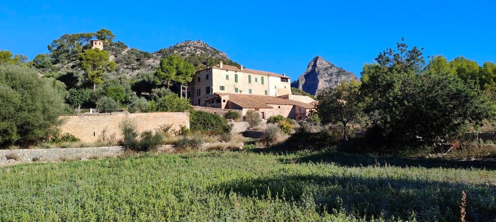 Vista de una finca rural situada en una llanura, rodeada de vegetación y montañas. Se aprecian casas de piedra con ventanas verdes, un pequeño edificio con cúpula, y un cielo despejado.