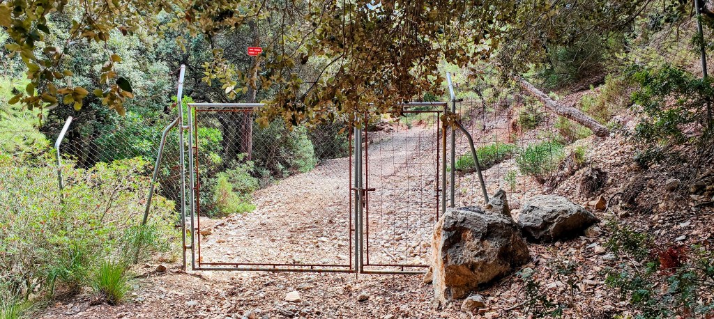 Puerta metálica cerrada en un camino de tierra, rodeada de vegetación densa y piedras.