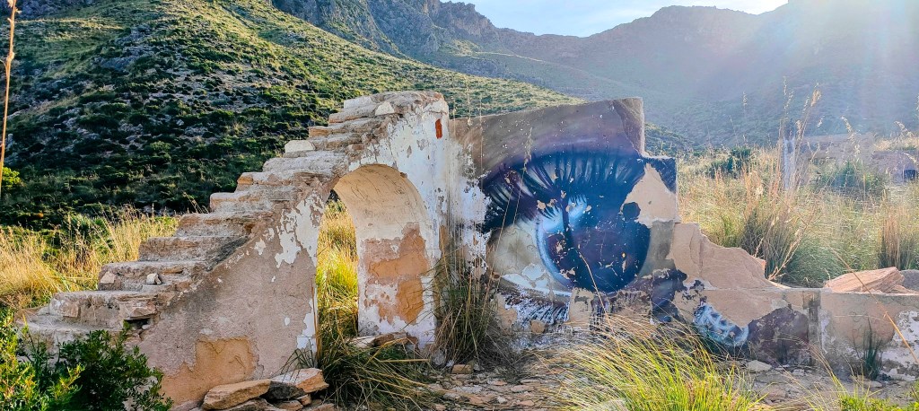 Muro en ruinas con una escalinata y un mural de un ojo, rodeado de vegetación y montañas al fondo.