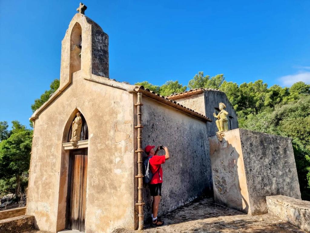 Vista exterior del Oratorio de Son Mascord, una pequeña ermita de montaña con figuras decorativas en su fachada, mientras un visitante toma una fotografía.