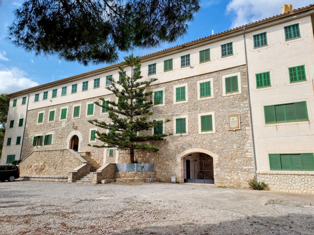 Edificio de piedra con ventanas verdes y escalones en la entrada, rodeado de árboles en un área de estacionamiento de grava.
