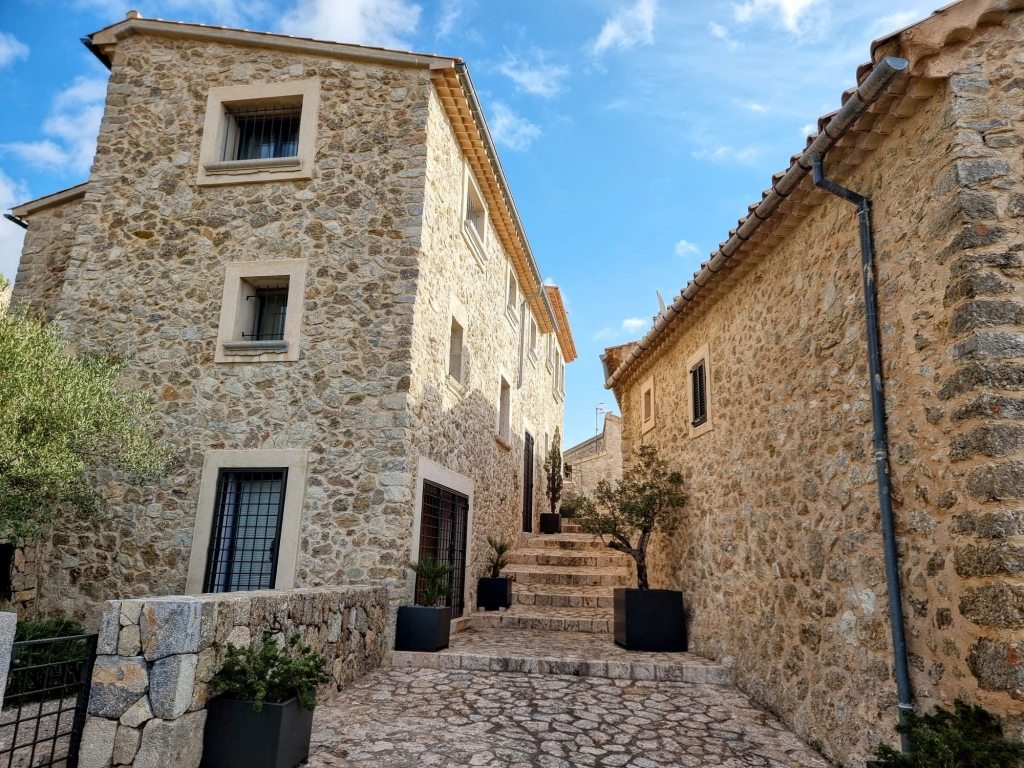 Vista de una calle de piedra con casas de piedra, plantas en macetas y un cielo parcialmente nublado.