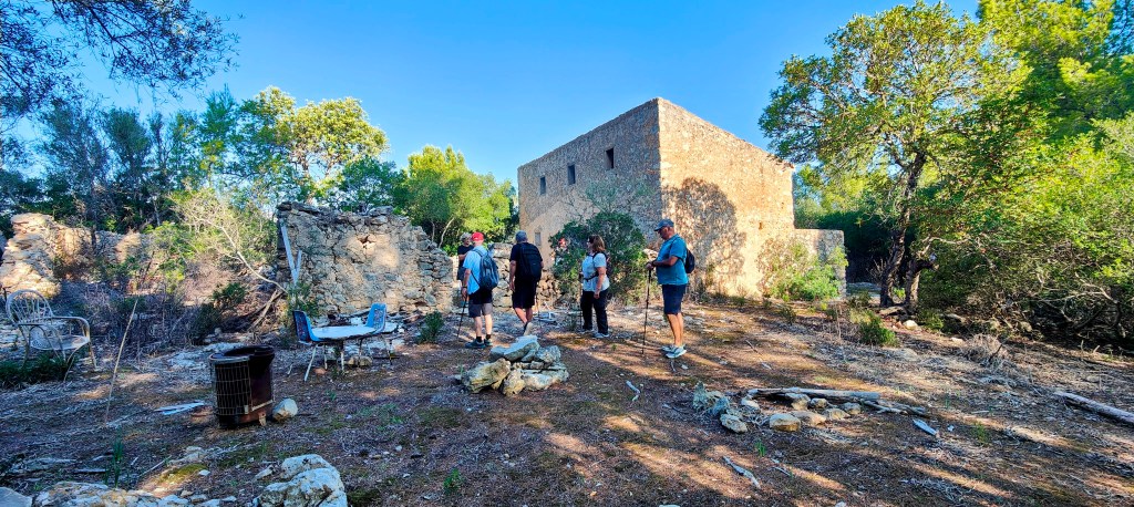 Grupo de personas explorando la zona de Son Mascord Vell en un paisaje montañoso con vegetación. Se pueden ver restos de una construcción antigua y árboles alrededor.