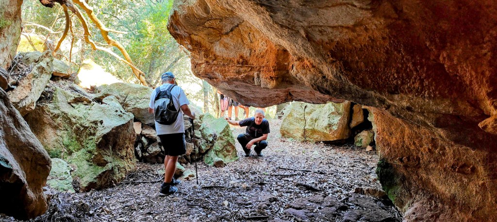 Dos personas explorando la Cueva de Baldritja bajo una gran roca, rodeados de vegetación y piedras.
