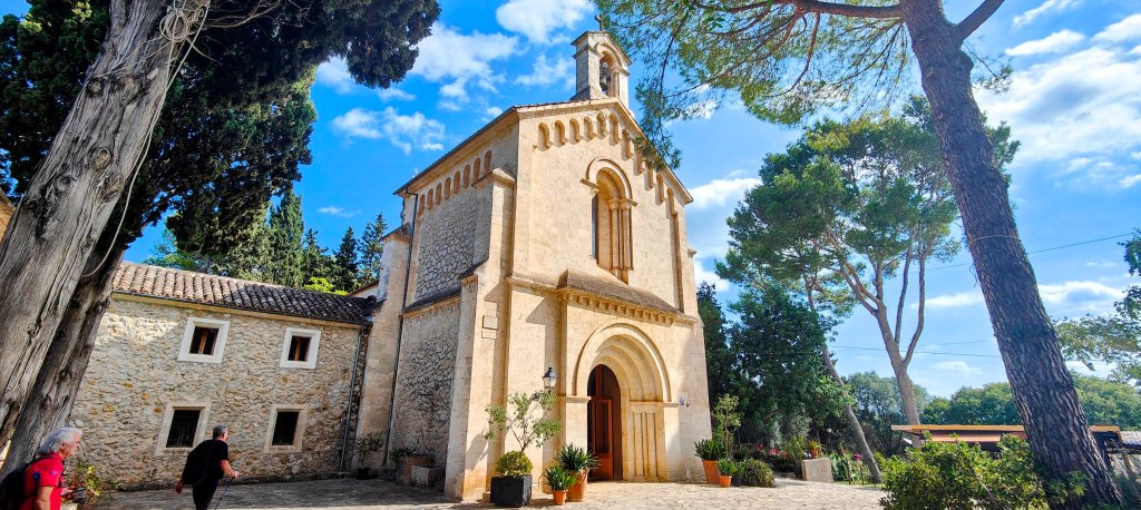 Vista de la Ermita de Crestatx, un pequeño edificio religioso con fachada de piedra, rodeado de vegetación y árboles altos, donde se observan personas caminando cerca.